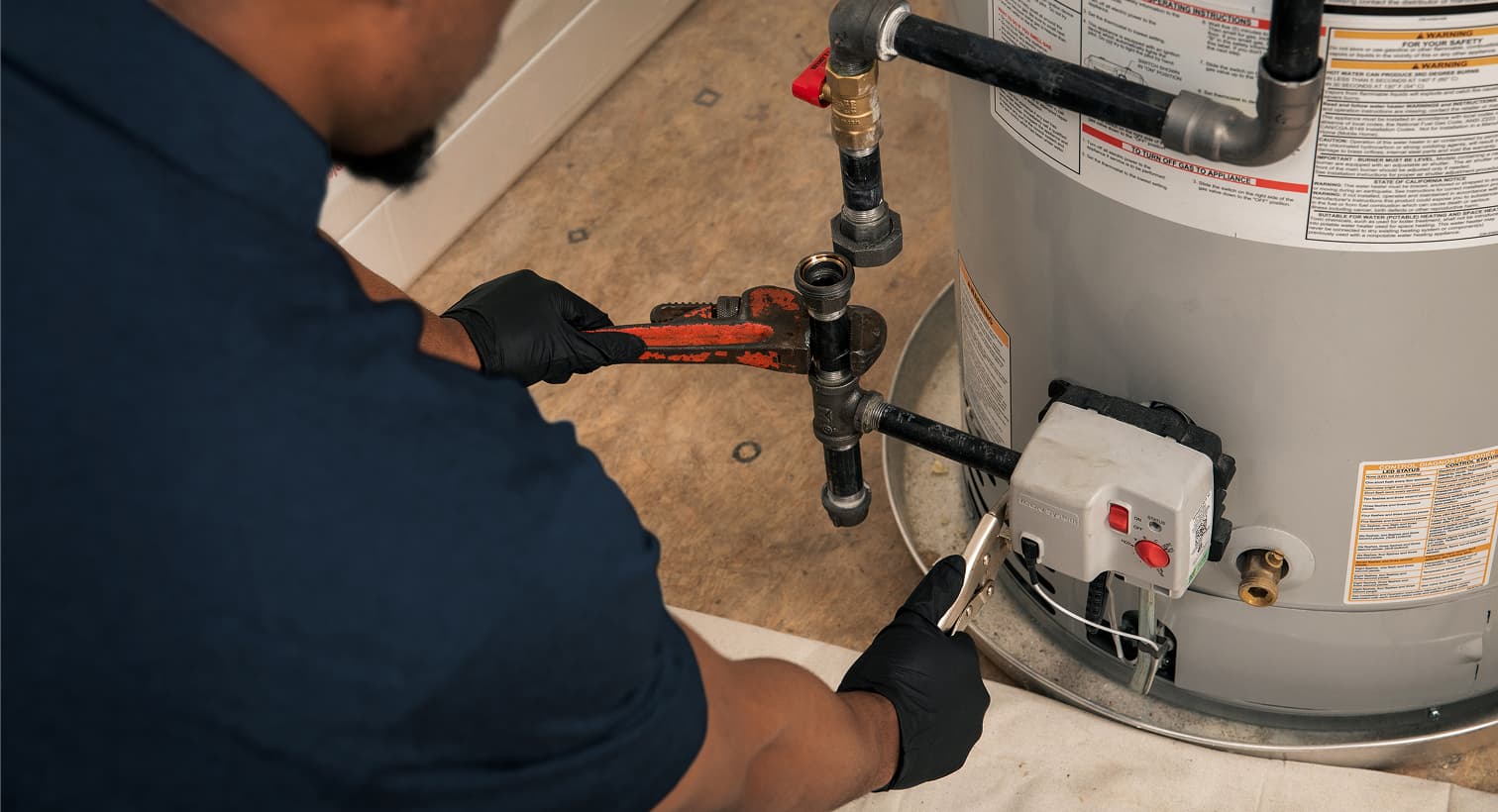 A Eanes plumber wearing black gloves uses a pipe wrench to repair the gas line and control valve assembly on a residential water heater tank.